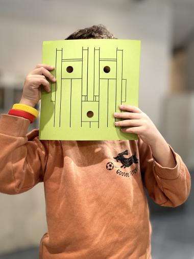 bambino con un cartoncino davanti alla faccia con un disegno di un volto fatto a righe geometriche